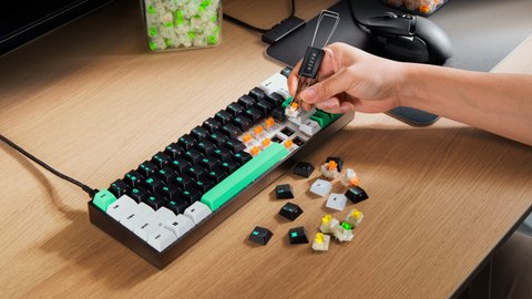 Una mano extrae un switch del teclado con una pinza extractora, con switches y teclas sueltas sobre la mesa